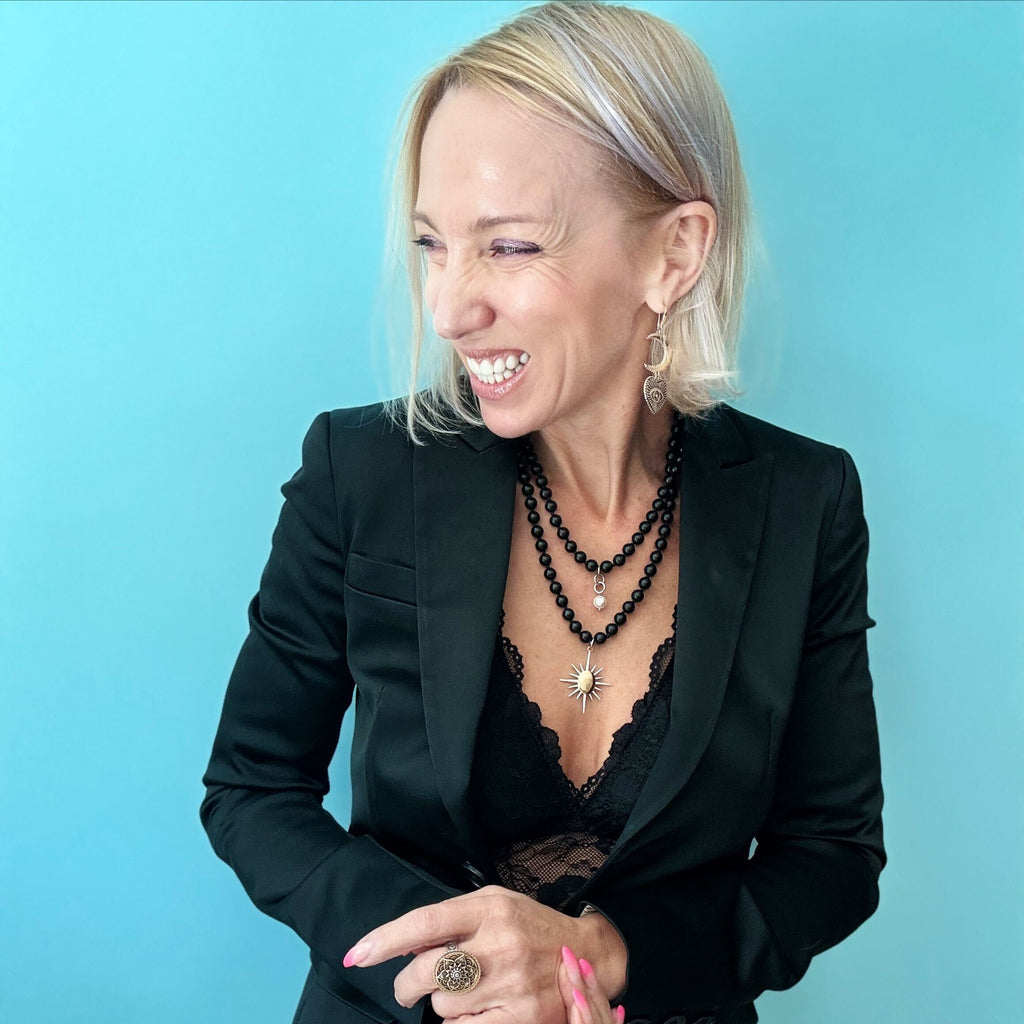 A woman in a black lace top and blazer stands against a blue background, her look elevated by a striking sunburst pendant.