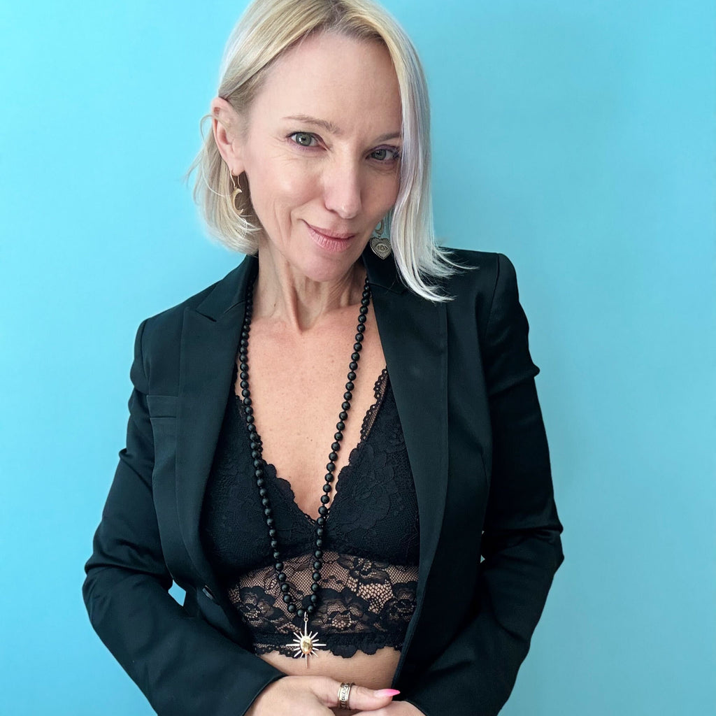 A woman in a black lace top and blazer stands against a blue background, her look elevated by a striking sunburst pendant.