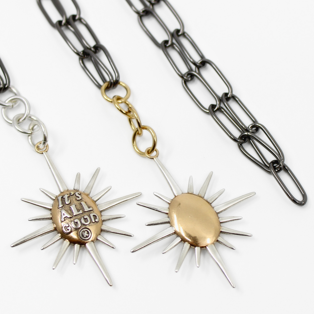 sunburst pendants on chains; one says Its all good and features a mix of silver and gold tones. both on a sleek gunmetal chain, offering a modern twist with its subtle magnetic clasp for easy wear.