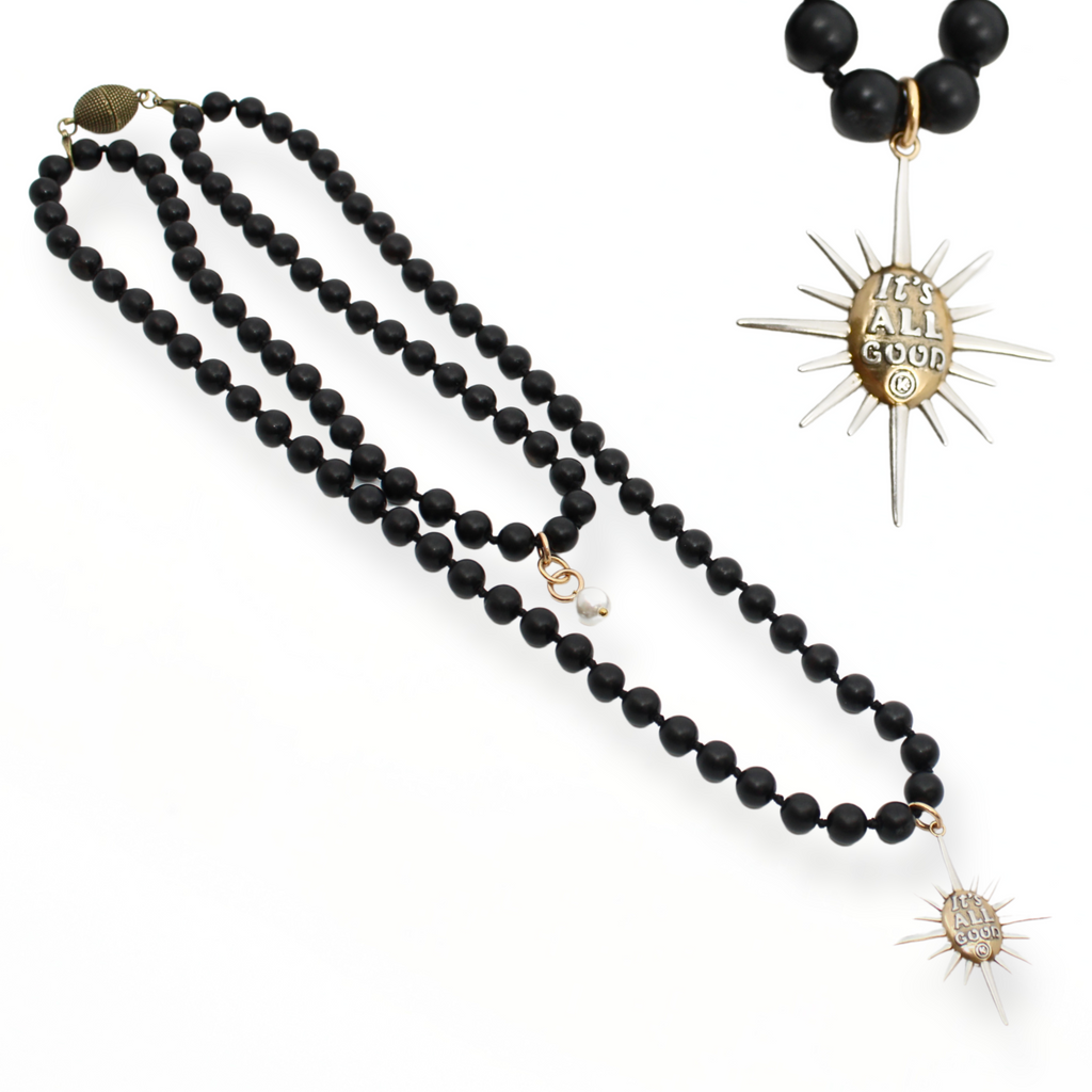 Black onyx necklace with a sunburst pendant reading Its All Good. Gold clasp and pearl accent detail on chain.