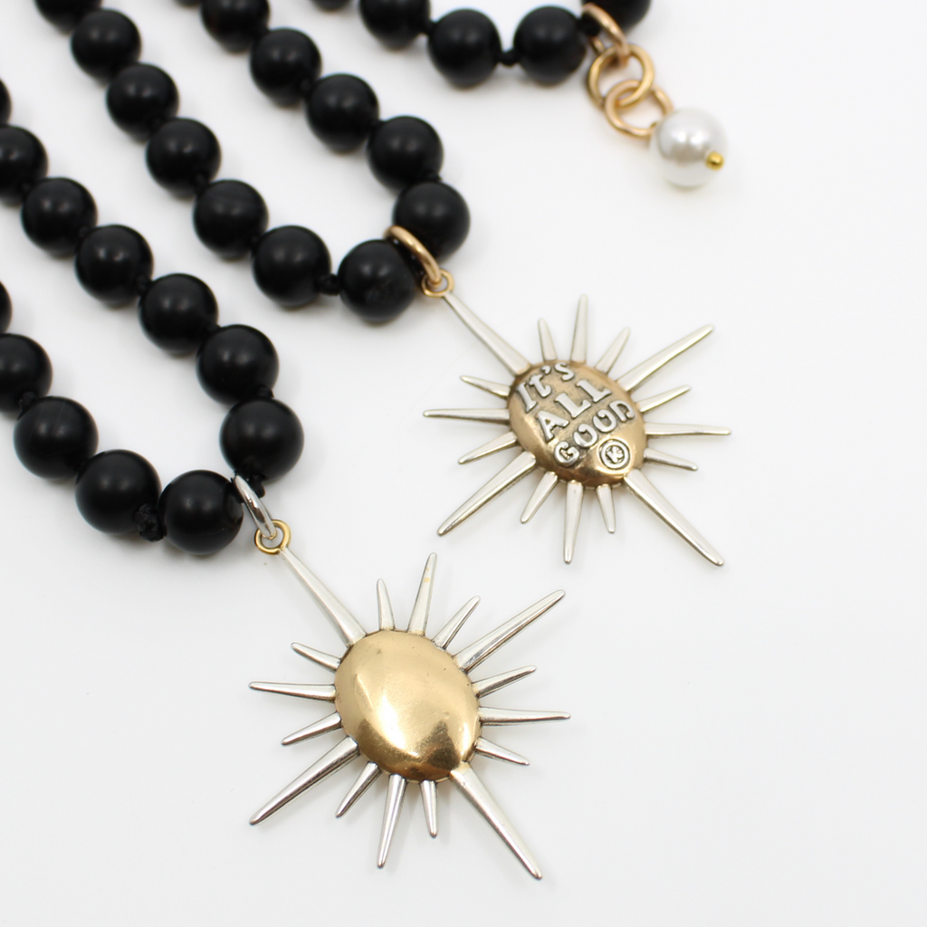 Black onyx necklace with a sunburst pendant reading Its All Good. Gold clasp and pearl accent detail on chain.