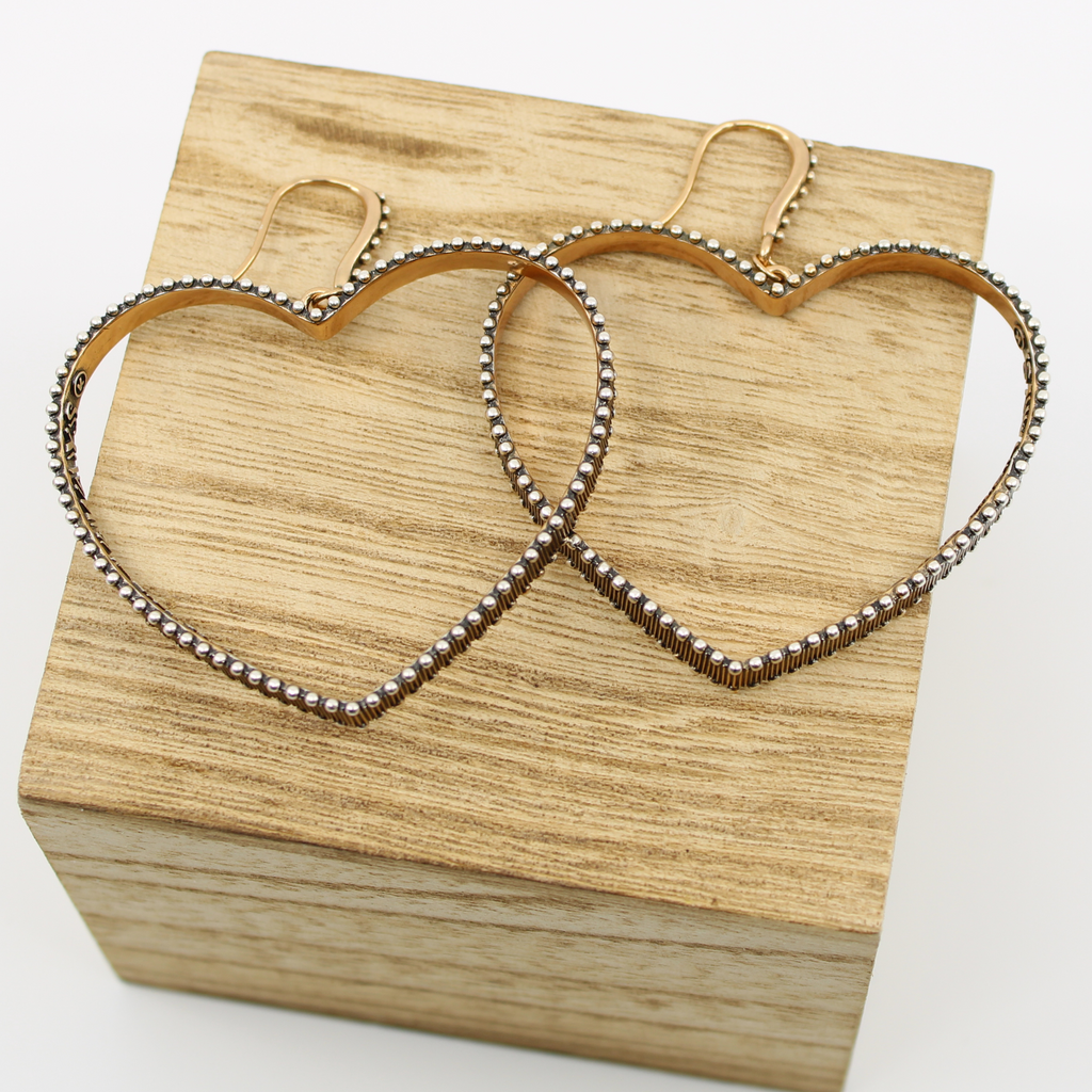 A pair of heart-shaped hoop earrings with textured edges and a mixed metal design sit on a wooden box. The earrings are metallic with French ear wire clasps at the top, contrasting elegantly against the plain white background.