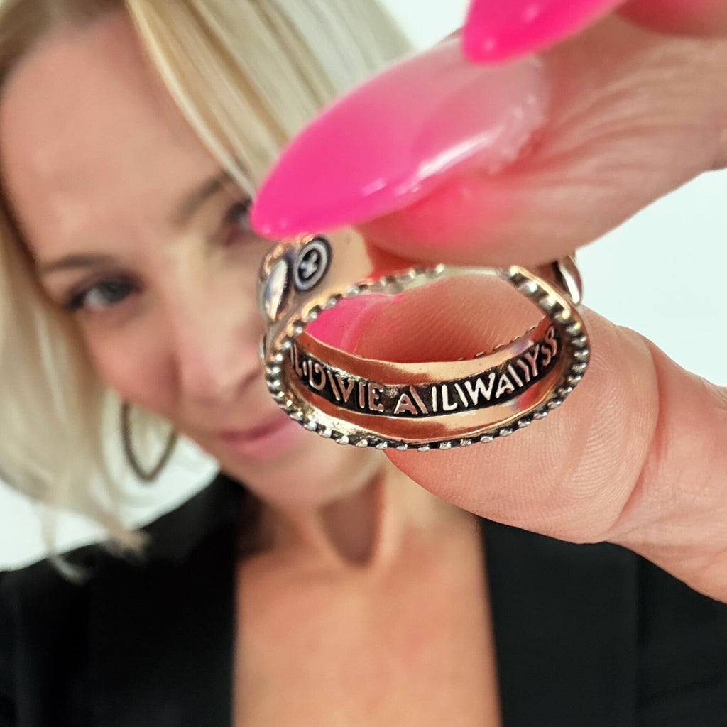 KATIA DESIGNS HOLDING A NEW mixed-metal ring close to the camera, showcasing the engraved words LOVE ALWAYS. They are wearing a black top, and their nails are painted bright pink, complementing the raised heart design on the adjustable band. The background is beautifully blurred.