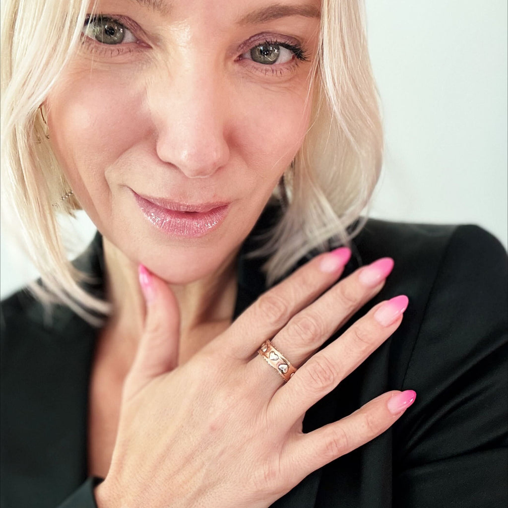 KATIA DESIGNS smiles gently, SHOWS OFF pink nail polish and hold their hand to their chest, displaying a mixed-metal ring with an adjustable band adorned in a small heart design. The background is plain and light-colored.