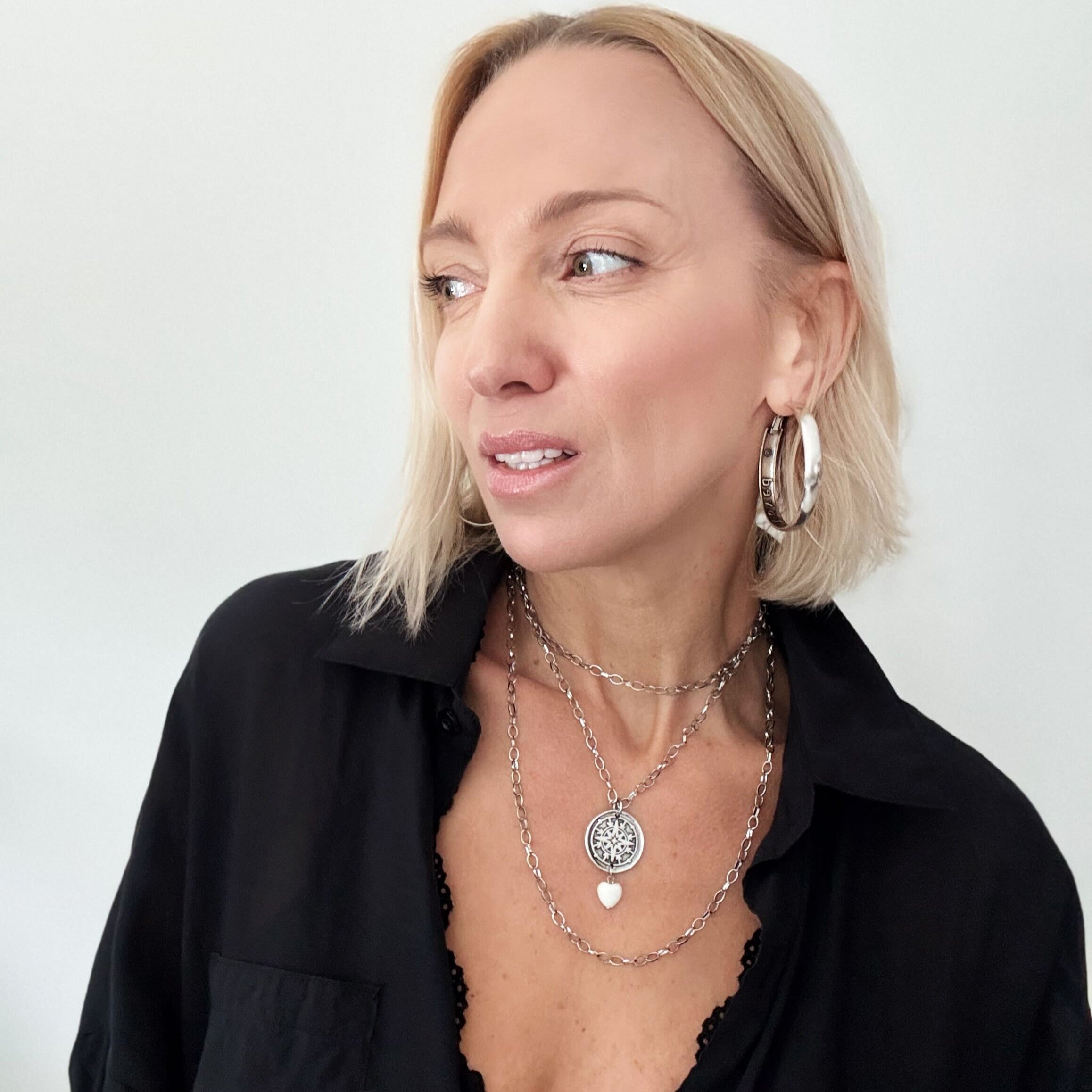 A smiling person with blonde hair wears a black shirt and layered necklaces, including a delicate layering chain adorned with howlite heart beads, against a plain background.