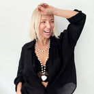 A smiling person with blonde hair wears a black shirt and layered necklaces, including a delicate layering chain adorned with howlite heart beads, against a plain background.