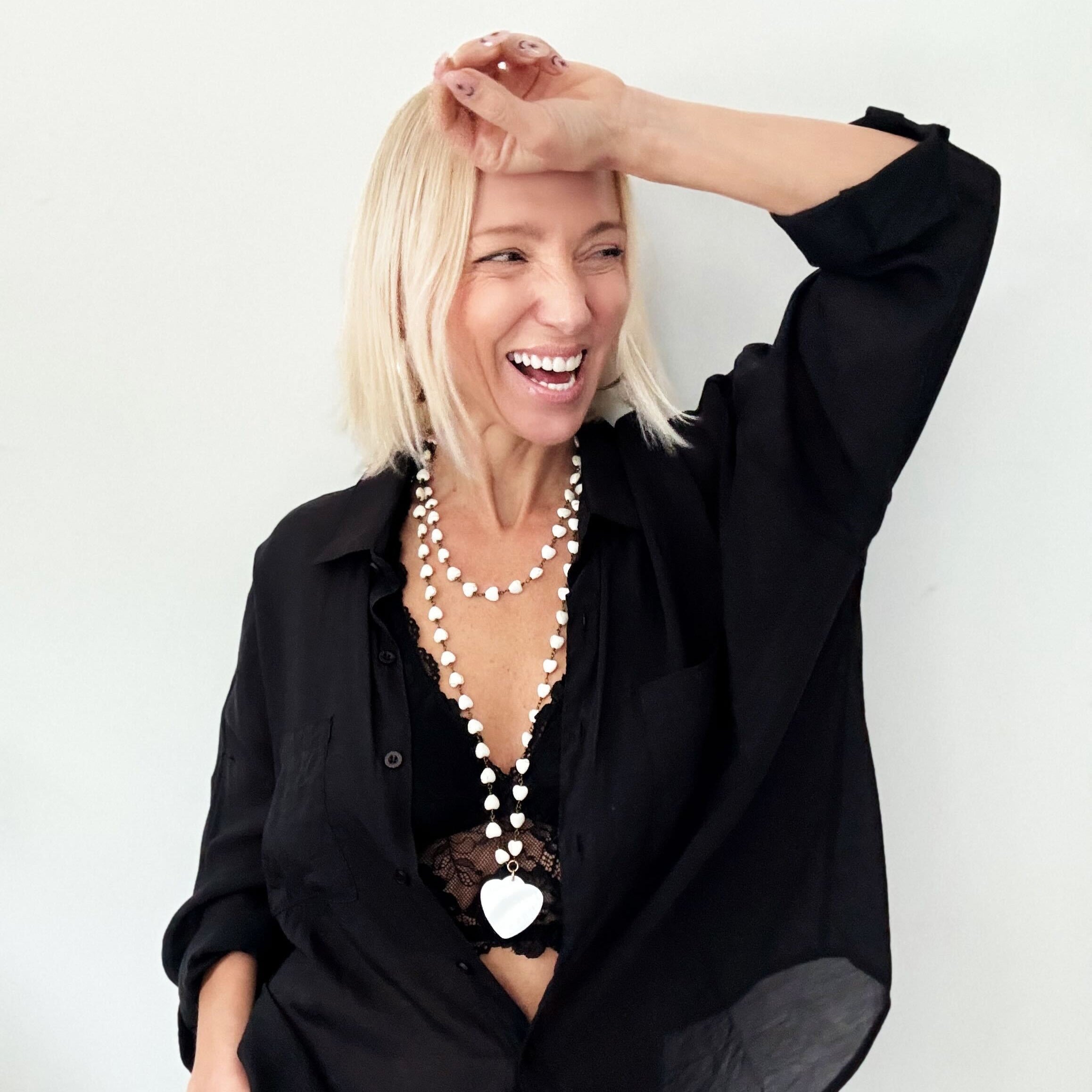 A smiling person with blonde hair wears a black shirt and layered necklaces, including a delicate layering chain adorned with howlite heart beads, against a plain background.