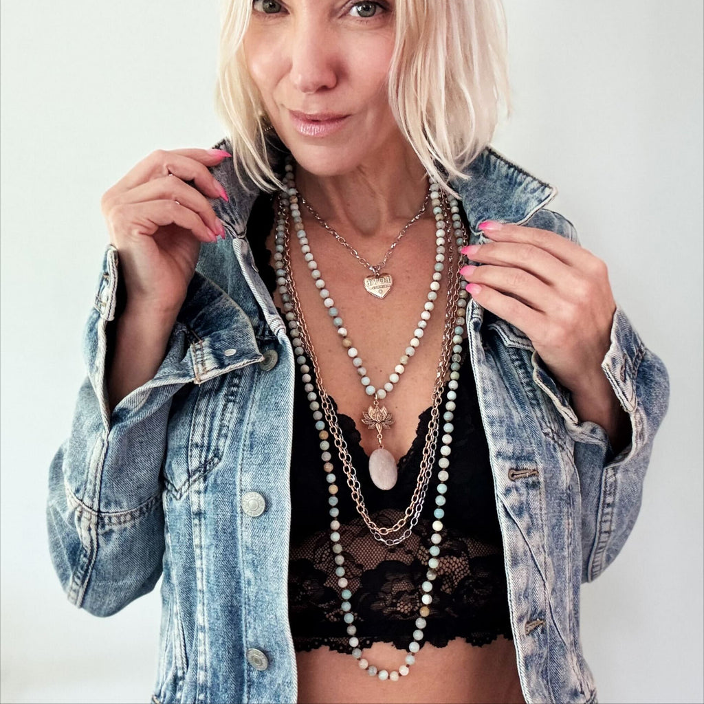 A person with short blond hair is wearing a denim jacket over a black top, accessorized with layered gold necklaces featuring a heart pendant and chain earrings. The mixed metal pendant glints under the light as they smile, holding the jacket lapels.