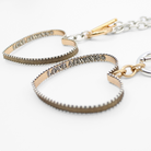 Close-up of two heart-shaped PENDANTS with textured edges, connected by a toggle closure. Each heart bears the inscription LOVE ALWAYS. They are placed on a white background.