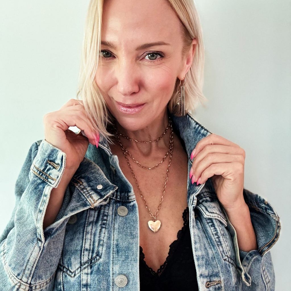 A person with short blond hair is wearing a denim jacket over a black top, accessorized with layered gold necklaces featuring a heart pendant and chain earrings. The mixed metal pendant glints under the light as they smile, holding the jacket lapels.