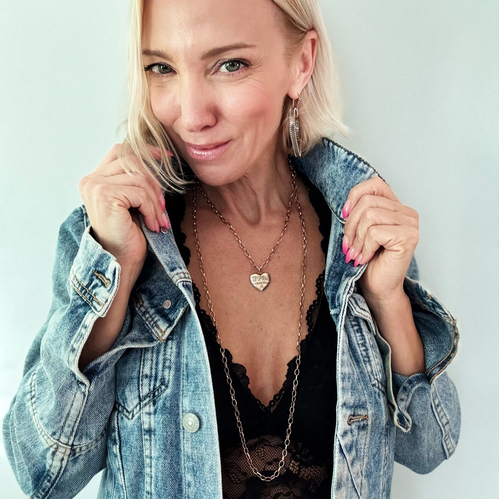 A person with short blond hair is wearing a denim jacket over a black top, accessorized with layered gold necklaces featuring a heart pendant and chain earrings. The mixed metal pendant glints under the light as they smile, holding the jacket lapels.