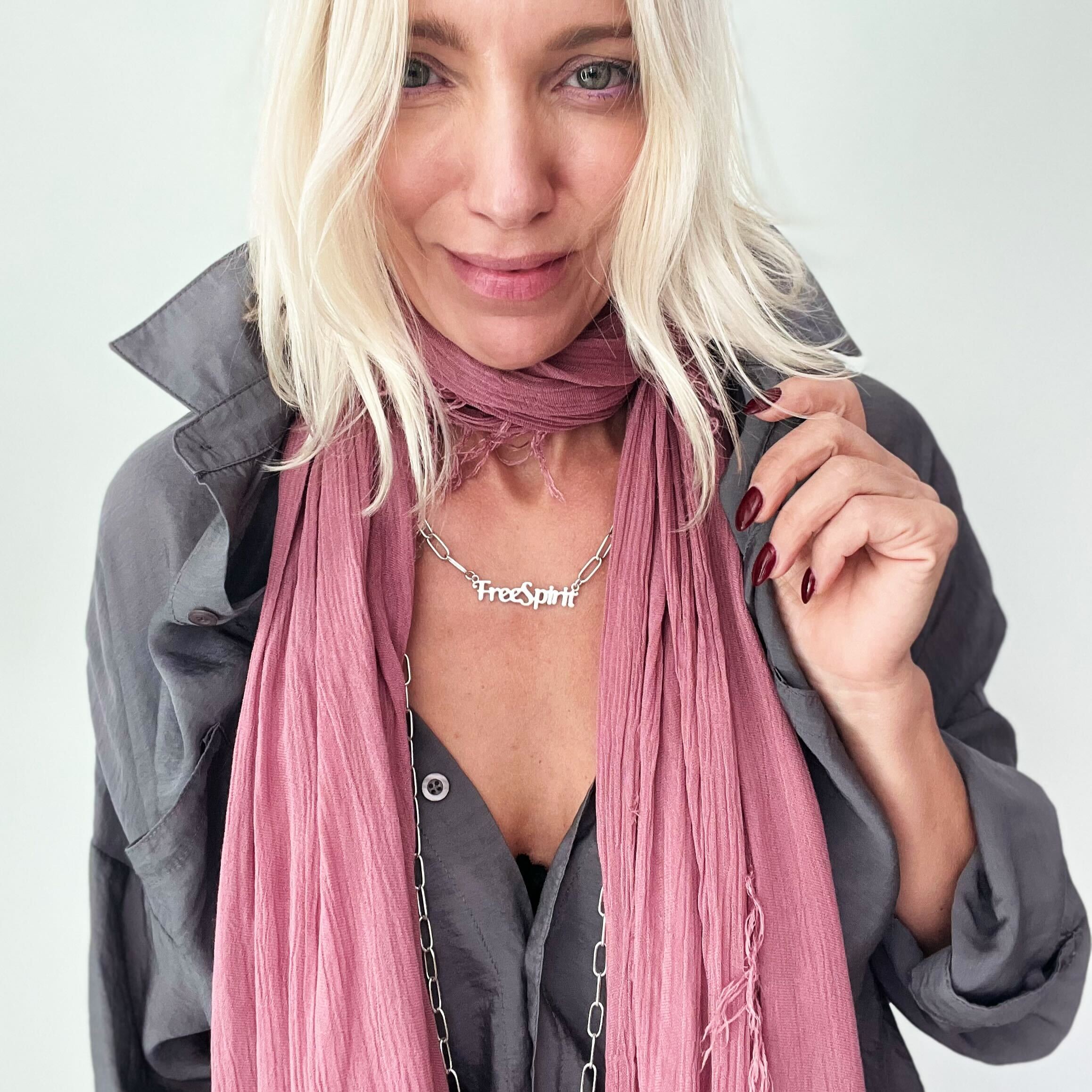 A person wearing a gray jacket and a pink scarf, showcasing a silver-colored long chain necklace with 'Free Spirit' text.