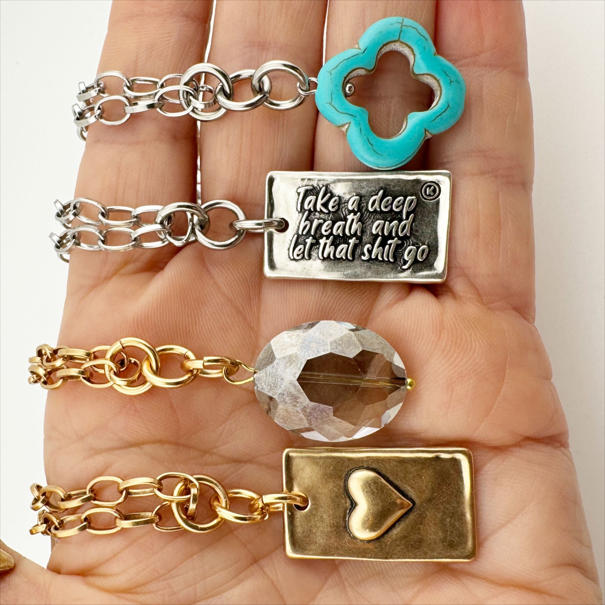 A collection of stainless steel necklaces with various pendants, including a turquoise clover, a clear faceted crystal, and a gold heart charm, held in a hand.