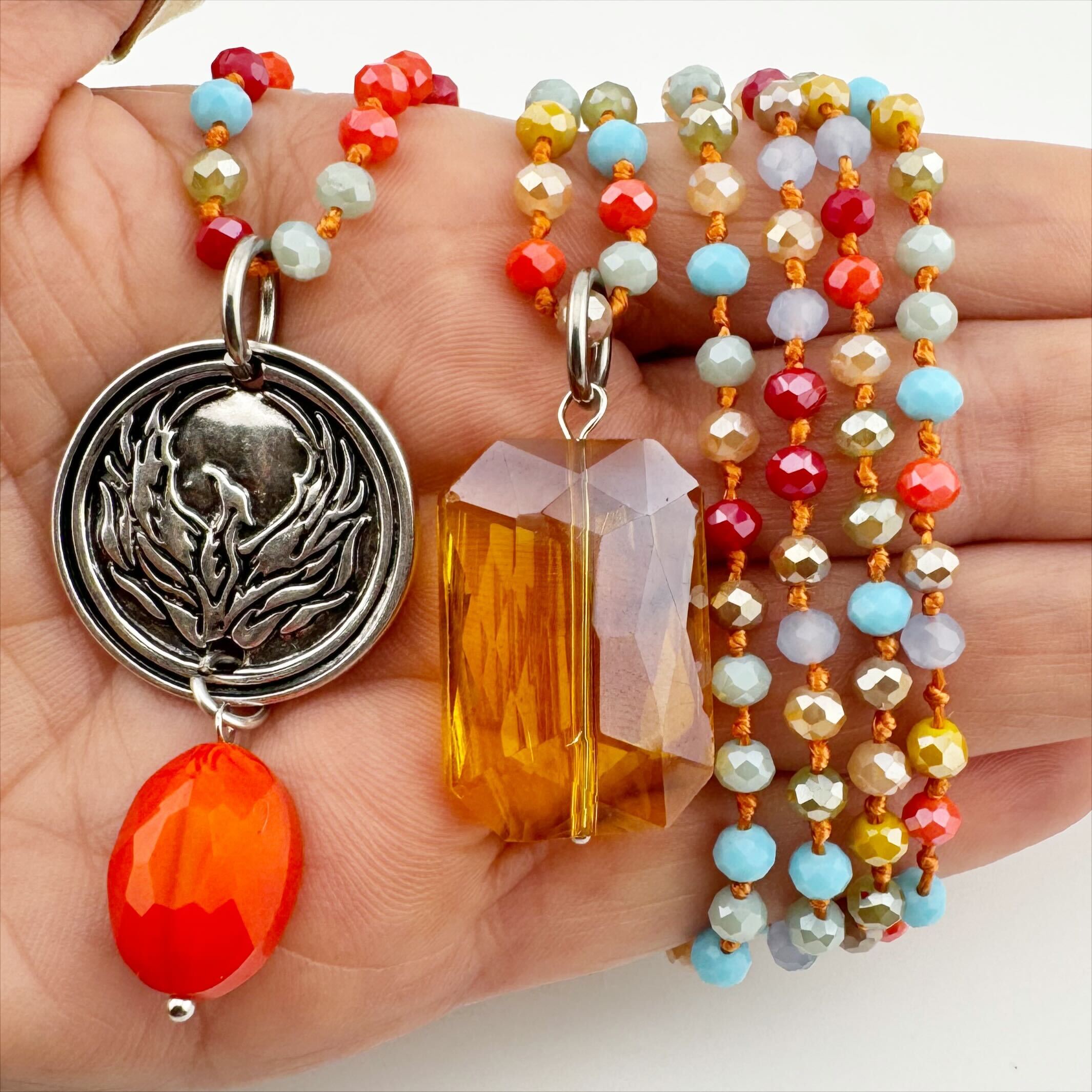 A person wearing a pink scarf and multiple necklaces with a variety of beads, including multicolored crystals and a gold-finished chain with a rectangular gunmetal crystal.