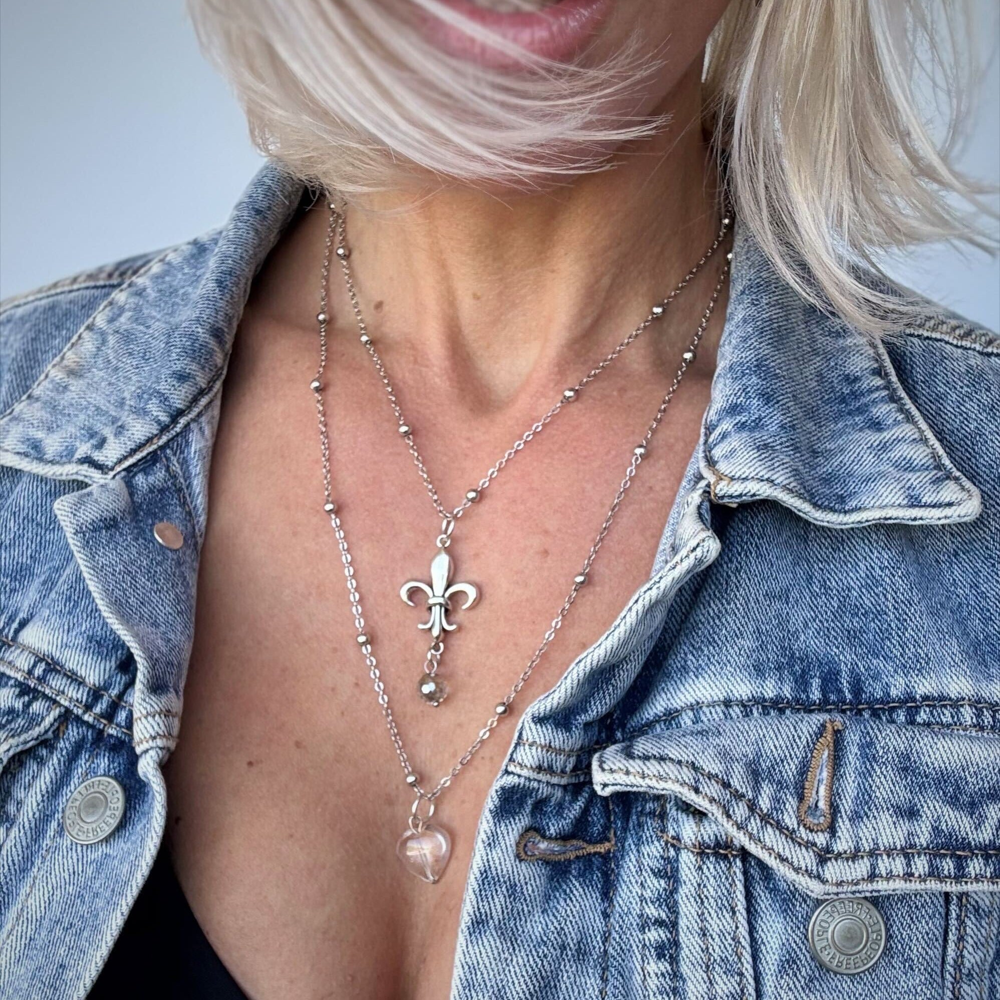 Person wearing a denim jacket with layered necklaces featuring a fleur-de-lis pendant.