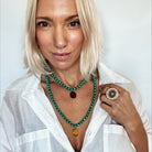 Woman wearing a white shirt and green beaded necklace with a large ring, against a plain background.