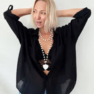 A smiling person with blonde hair wears a black shirt and layered necklaces, including a delicate layering chain adorned with howlite heart beads, against a plain background.