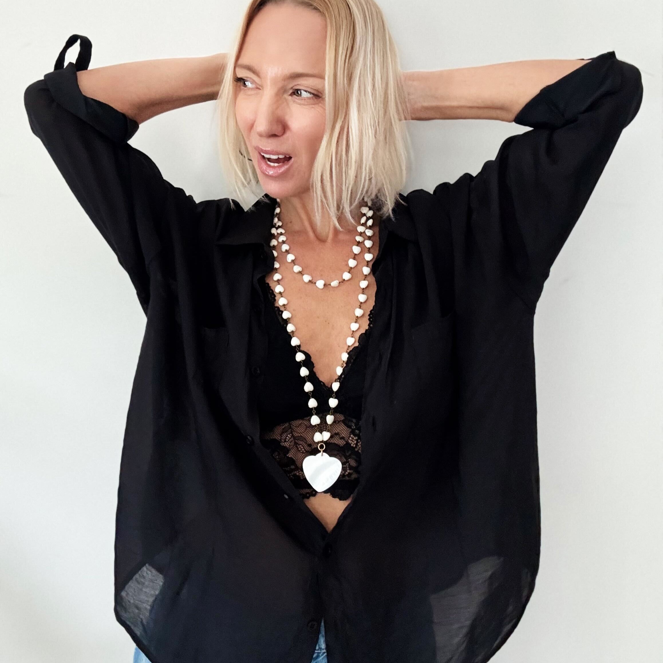 A smiling person with blonde hair wears a black shirt and layered necklaces, including a delicate layering chain adorned with howlite heart beads, against a plain background.