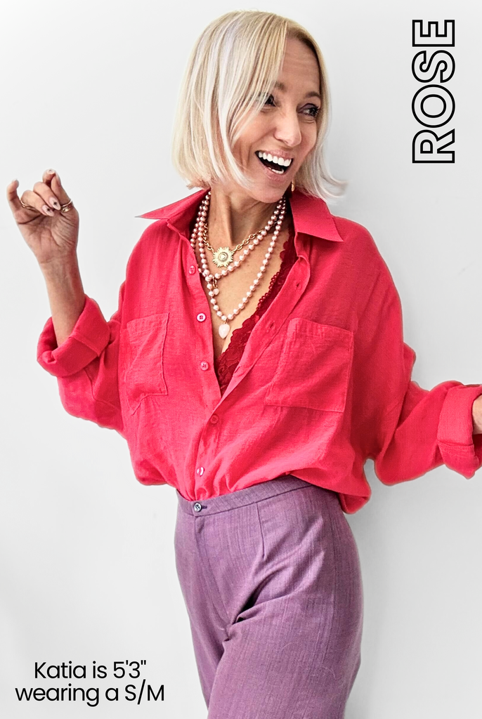 Woman wearing a bright pink, boho-chic shirt with layered necklaces and purple pants smiles with a sideways glance. Text reads Katia is 53 wearing a S/M and ROSE.