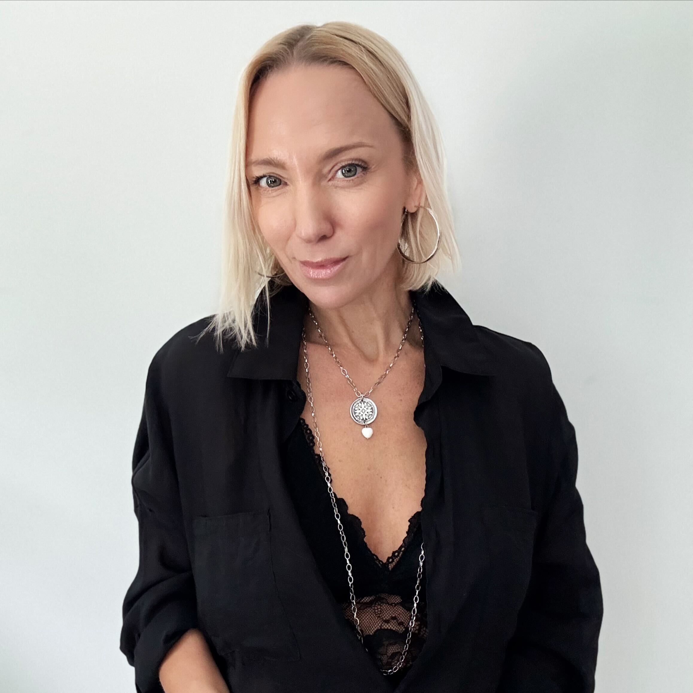A smiling person with blonde hair wears a black shirt and layered necklaces, including a delicate layering chain adorned with howlite heart beads, against a plain background.
