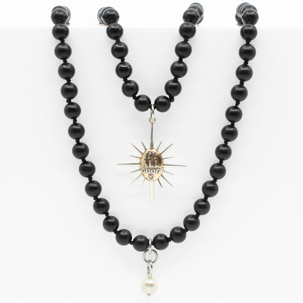 Black onyx necklace with a sunburst pendant reading Its All Good. Gold clasp and pearl accent detail on chain.