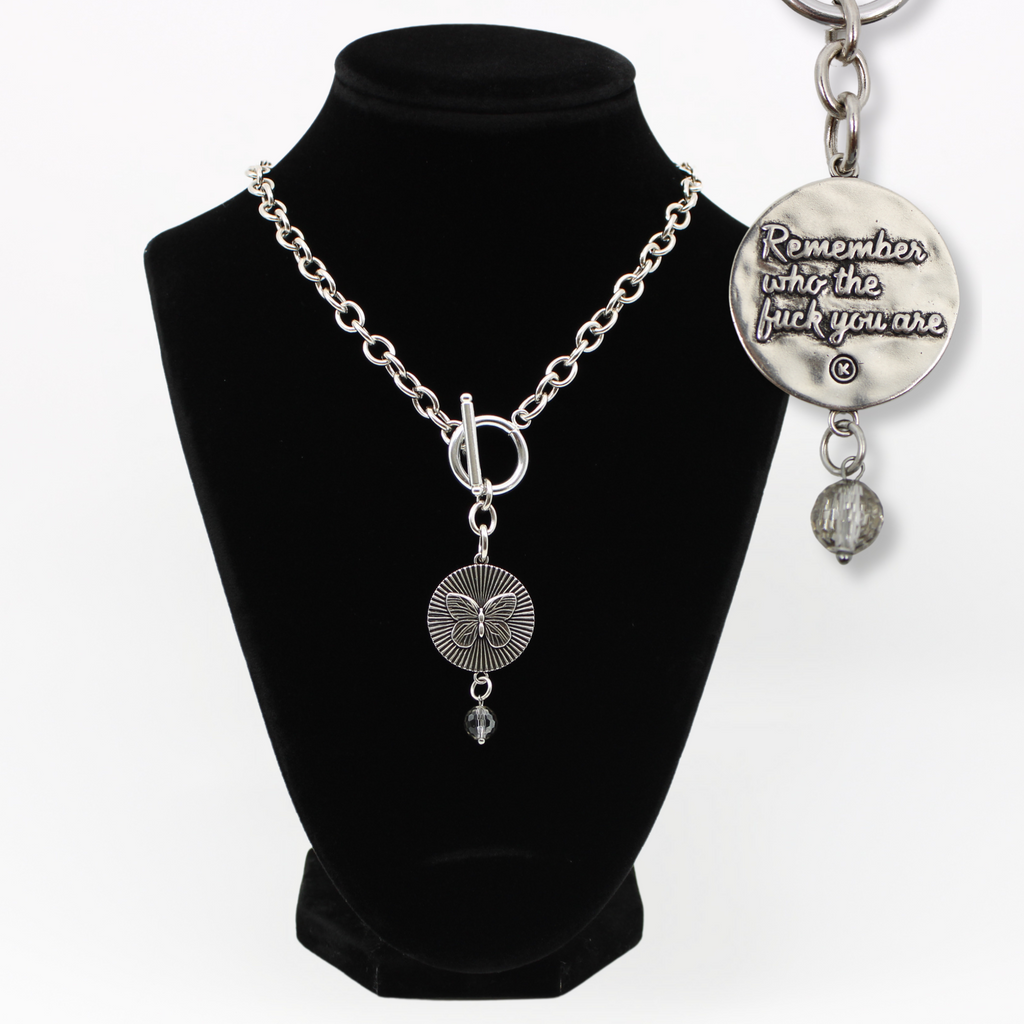 DETAILS AND PRODUCT DISPLAY OF SILVER VARIANT ON BUST; one with a butterfly pendant and crystal accent, the other boldly inscribed with Remember who the fuck you are.