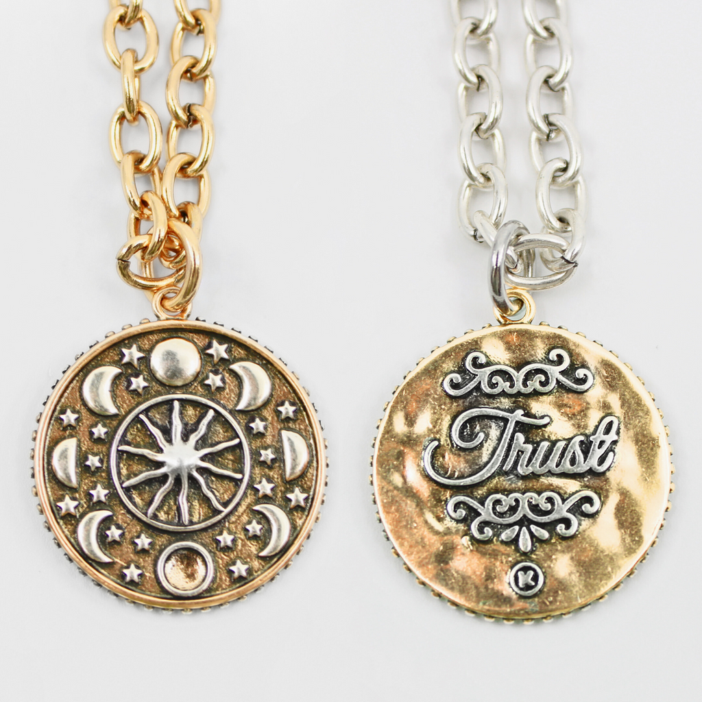 Two views of a decorative medallion necklace: front with moon phases, the other with the word Trust inscribed. Both feature a sleek magnetic clasp for easy wear and a mixed-metal pendant design that adds elegance.