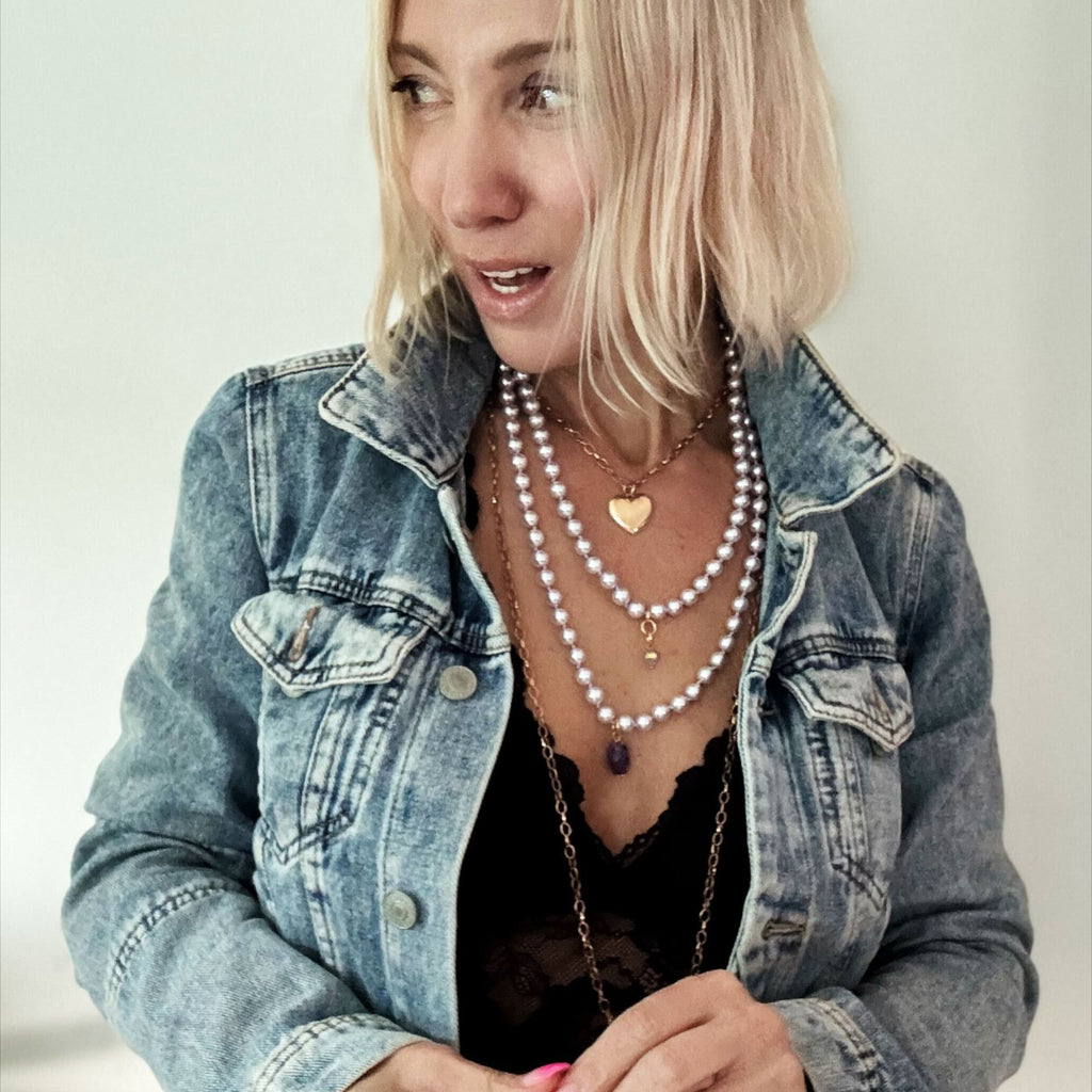 A person with short blond hair is wearing a denim jacket over a black top, accessorized with layered gold necklaces featuring a heart pendant and chain earrings. The mixed metal pendant glints under the light as they smile, holding the jacket lapels.