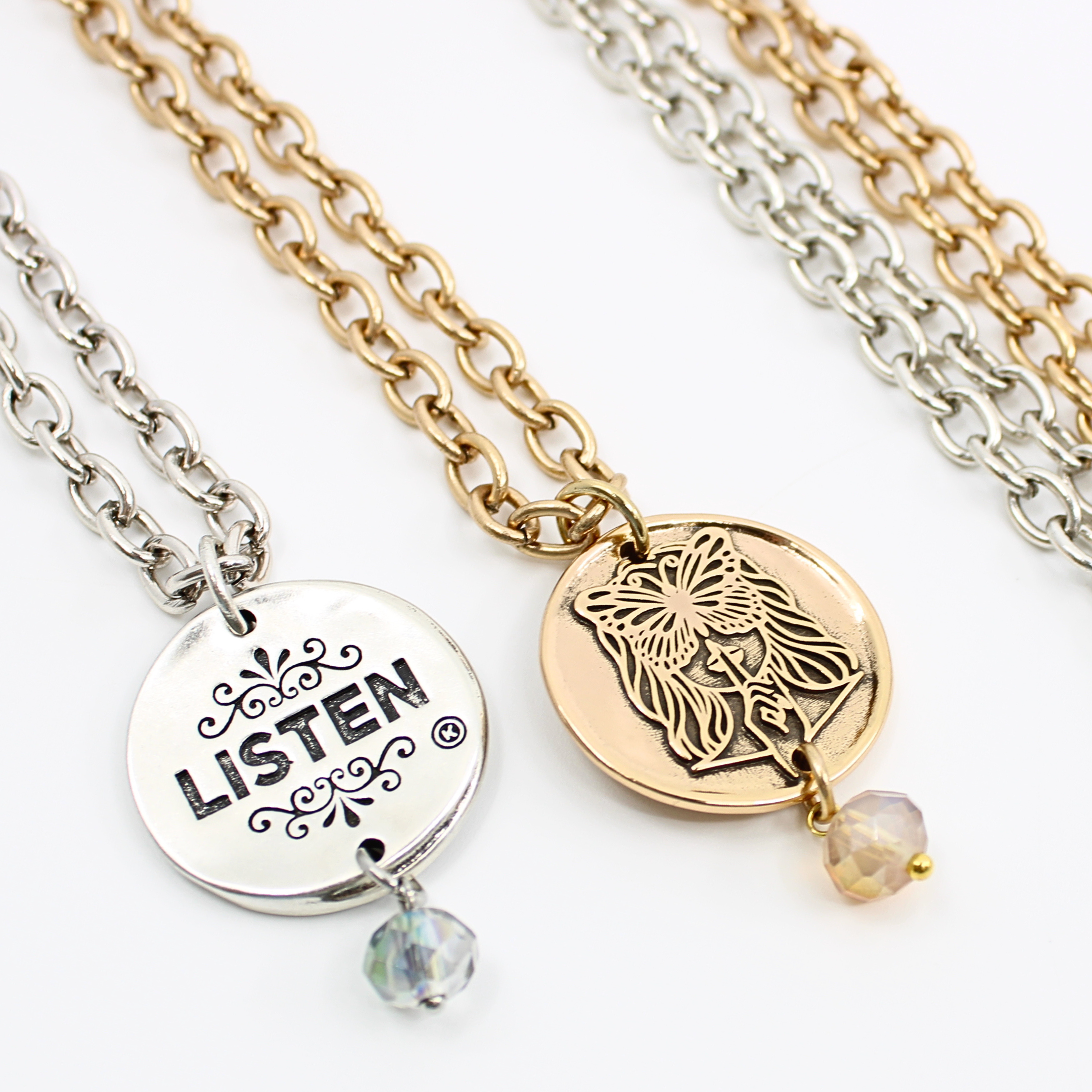 Two chain necklaces with round pendants: one silver with LISTEN adorned in plated brass and small beads, the other gold featuring a face design. Each piece exudes charm, enhanced by subtle crystal accents.