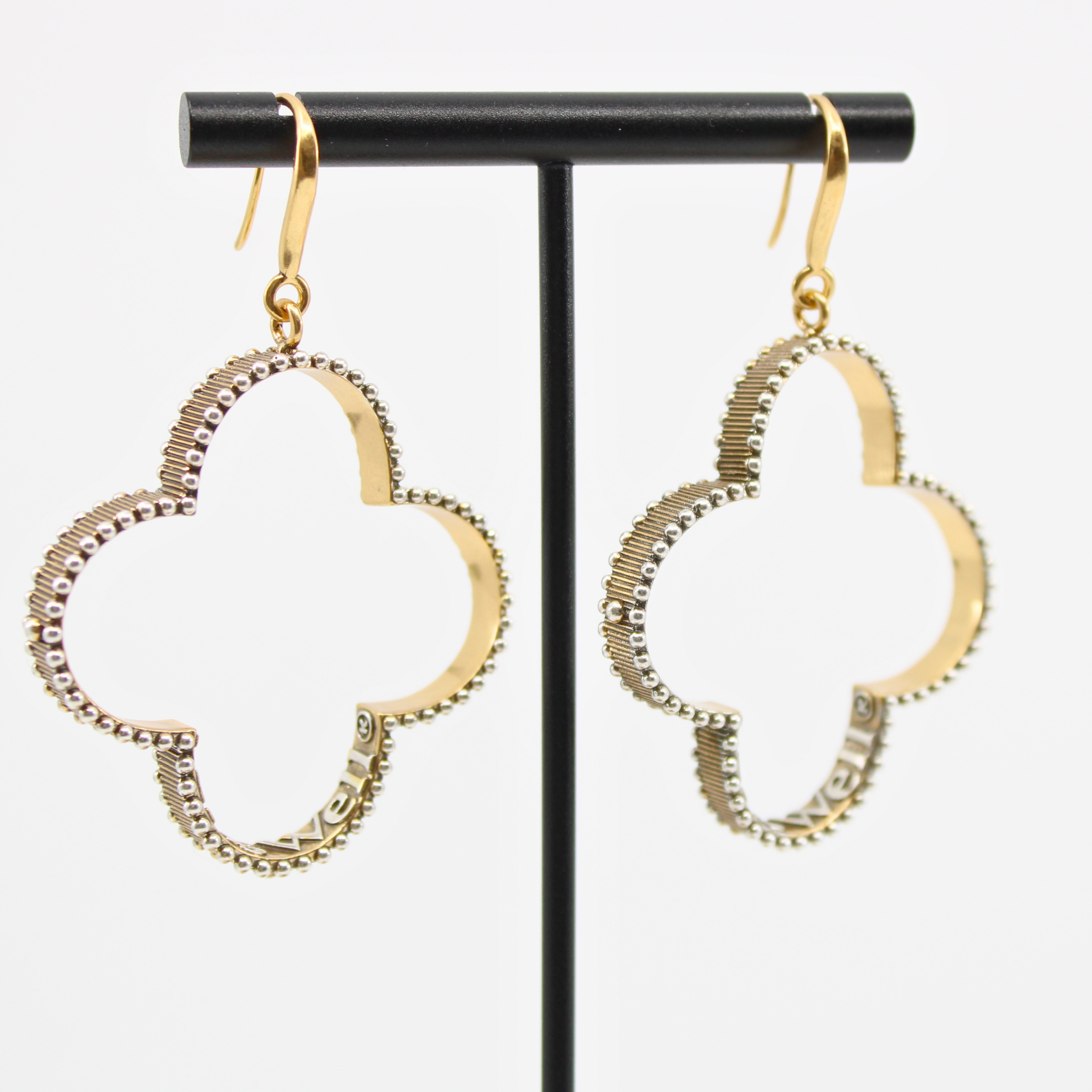 A pair of gold-colored composite drop earrings with French ear wire, featuring a clover design with a message inscribed inside, displayed on an earing stand.