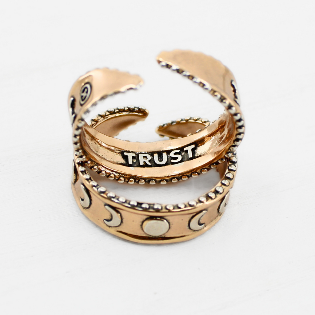 two mixed-metal rings with crescent moon designs and TRUST engraved inside, each featuring adjustable bands, are shown from different angles.