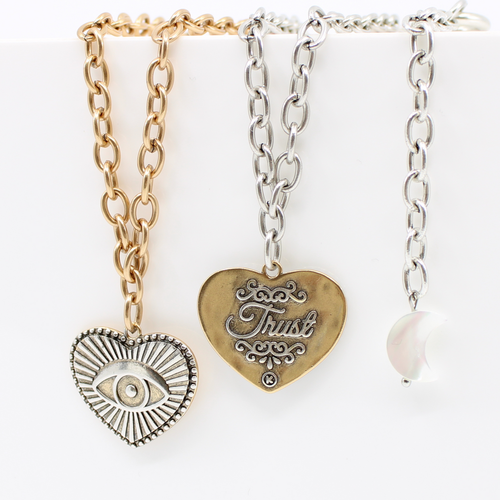 Gold or silver chain choker with a heart-shaped eye pendant, bar clasp, and crescent moon charm. Its mixed metal design adds a unique flair, all set against a white background.