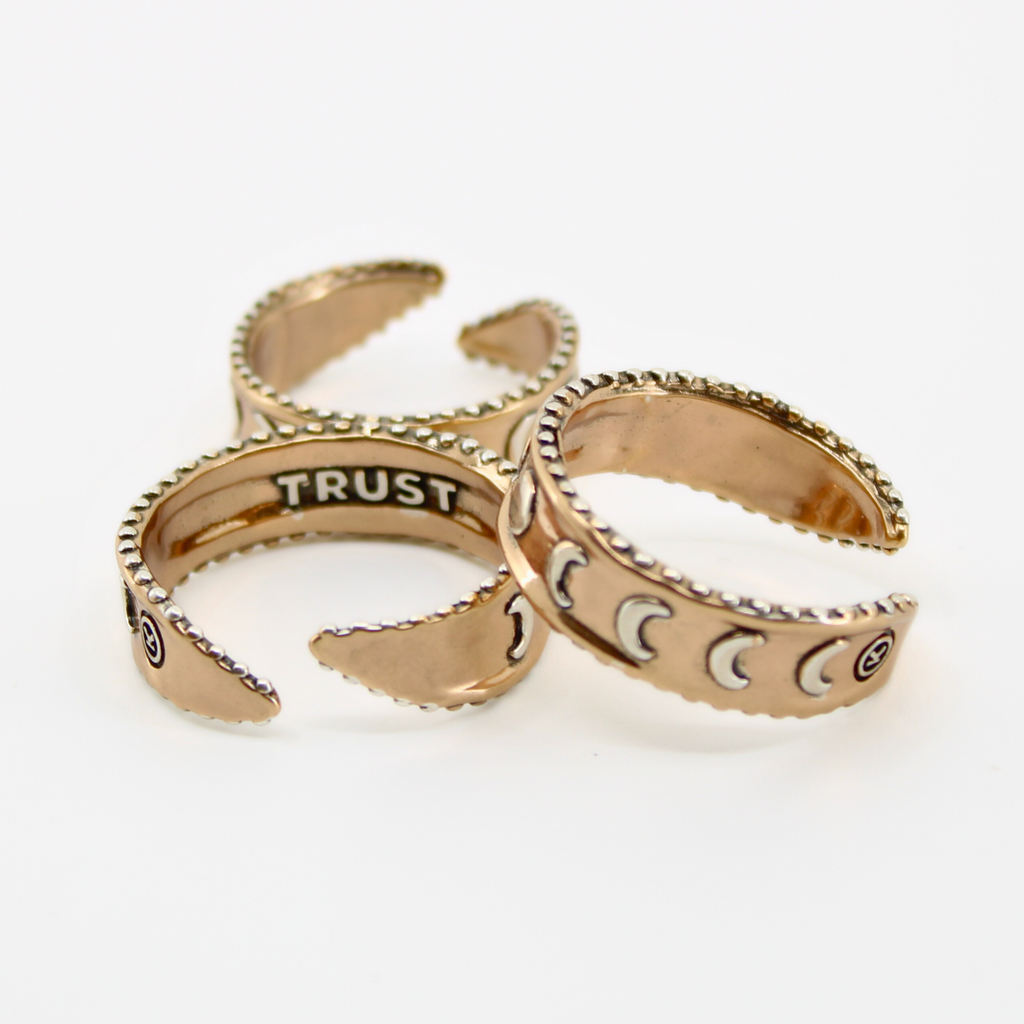 Three mixed-metal rings with crescent moon designs and TRUST engraved inside, each featuring adjustable bands, are shown from different angles.