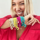 A person grins broadly, wearing a pink shirt and proudly showcasing multiple colorful beaded bracelets on their arm, one of which matches their boho wrap necklace adorned with a striking howlite beads.