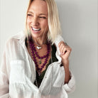 A person with blonde hair dons layered necklaces, showcasing a magenta purple howlite bead necklace and the heart locket essential in silver over a dark green top paired with a white shirt.