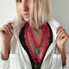 A person with blonde hair dons layered necklaces, showcasing a hot pink howlite bead necklace and a metallic "K" pendant, over a dark green top paired with a white shirt.