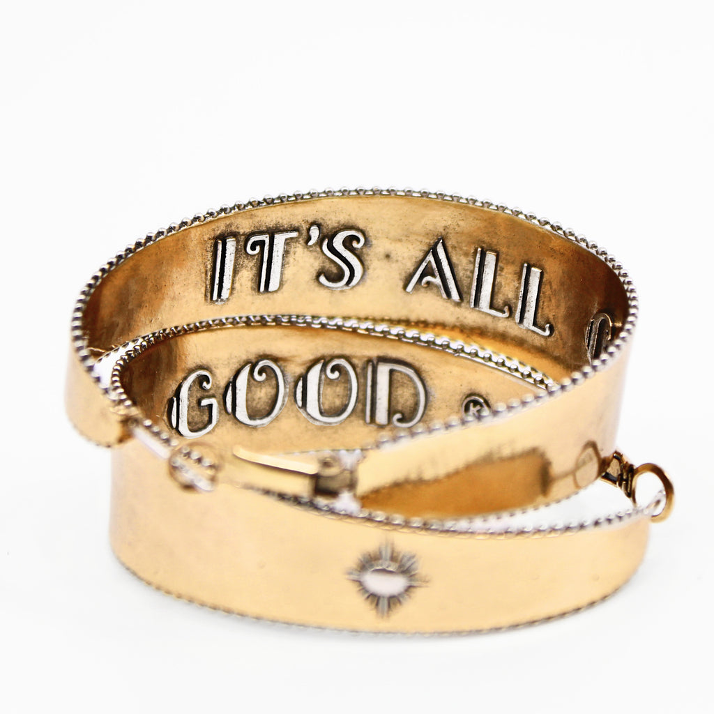 A pair of two-toned hoop earrings with intricate detailing. The outer surface features small, engraved sunburst designs with a tiny central accent. The edges of the hoops have a textured trim, adding to a vintage-inspired boho aesthetic. The inner band of the hoops is inscribed with raised lettering with the message "It's All Good." The earrings have a polished gold finish, giving them a stylish and elegant look.