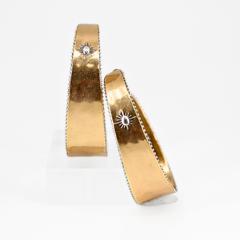 A pair of two-toned hoop earrings with intricate detailing. The outer surface features small, engraved sunburst designs with a tiny central accent. The edges of the hoops have a textured trim, adding to a vintage-inspired boho aesthetic. The inner band of the hoops is inscribed with raised lettering with the message "It's All Good." The earrings have a polished gold finish, giving them a stylish and elegant look.