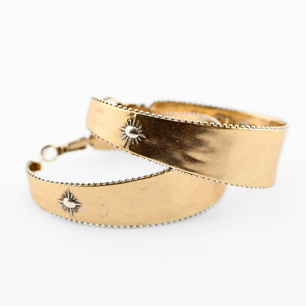 A pair of two-toned hoop earrings with intricate detailing. The outer surface features small, engraved sunburst designs with a tiny central accent. The edges of the hoops have a textured trim, adding to a vintage-inspired boho aesthetic. The inner band of the hoops is inscribed with raised lettering with the message "It's All Good." The earrings have a polished gold finish, giving them a stylish and elegant look.