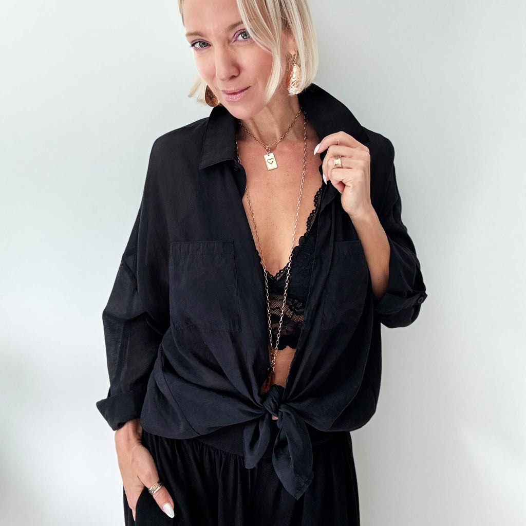 A person with blonde hair in a black top, adorned with a LOVE pendant and large earrings, is posing against a plain background.