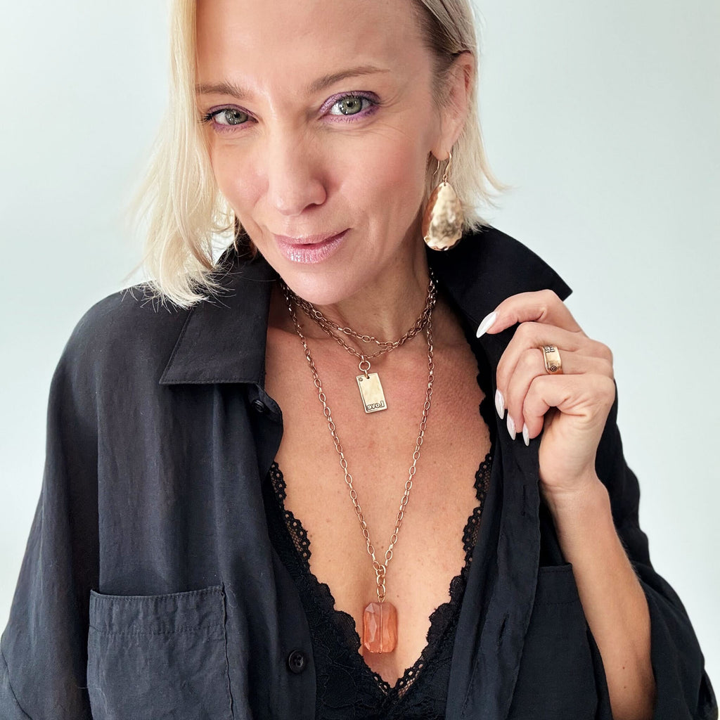 A person with blonde hair in a black top, adorned with a LOVE pendant and large earrings, is smiling against a plain background.