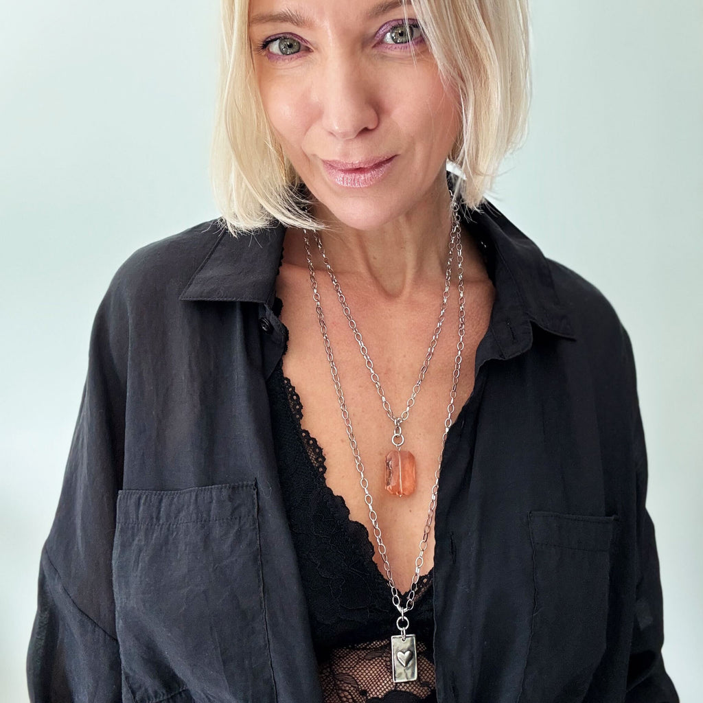 A person with blonde hair in a black top, adorned with a LOVE pendant and large earrings, is smiling against a plain background.