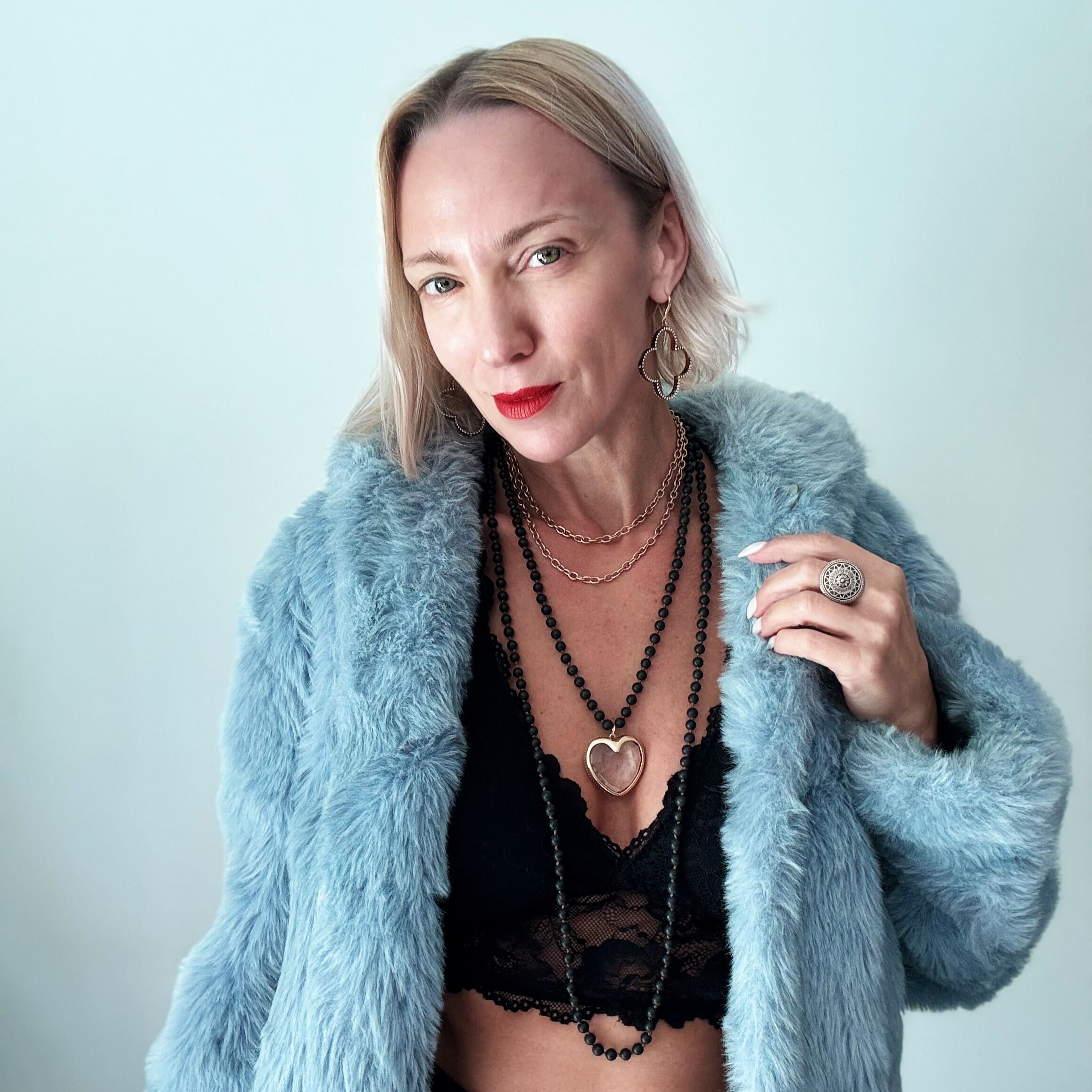 A person wearing a black lace top and a blue faux fur coat, showcasing a long black beaded necklace with a heart-shaped pendant.