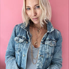 Person in a denim jacket and gray shirt poses against a pink background, showcasing layered necklaces with a chain necklace and rings shimmering with crystal accents.