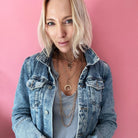Person in a denim jacket and gray shirt poses against a pink background, showcasing layered necklaces with a chain necklace and rings shimmering with crystal accents.