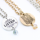 Two chain necklaces with round pendants: one silver with LISTEN adorned in plated brass and small beads, the other gold featuring a face design. Each piece exudes charm, enhanced by subtle crystal accents.