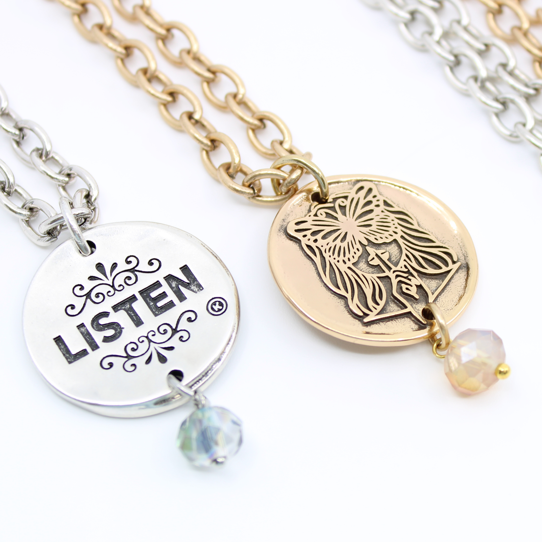 Two chain necklaces with round pendants: one silver with LISTEN adorned in plated brass and small beads, the other gold featuring a face design. Each piece exudes charm, enhanced by subtle crystal accents.