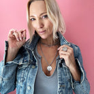 Person in a denim jacket and gray shirt poses against a pink background, showcasing layered necklaces with a chain necklace and rings shimmering with crystal accents.