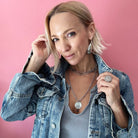 Person in a denim jacket and gray shirt poses against a pink background, showcasing layered necklaces with a chain necklace and rings shimmering with crystal accents.
