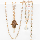 A collection of layered rosary necklaces with beige crystal beads, metallic chains, and decorative pendants including a peace symbol and a hand of Hamsa.